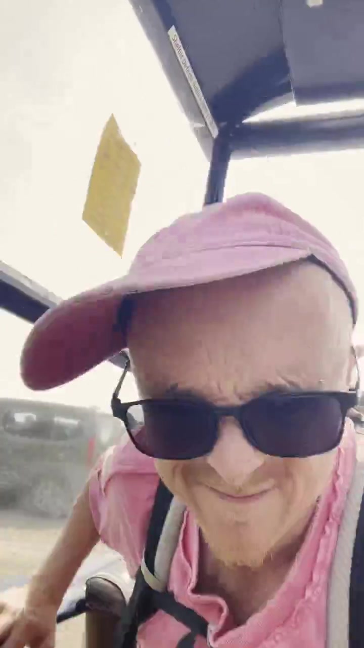 A vertical still of Gingerbeard in a pink cap and dark glasses, speaking in a sunlit Glasgow shelter.